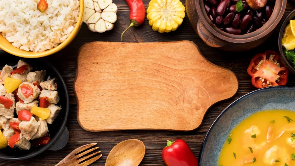 Mesa rústica de madeira com tábua central vazia cercada por arroz, feijão, milho, pimenta, alho, carne com legumes e caldo amarelo, representando comidas do sítio.