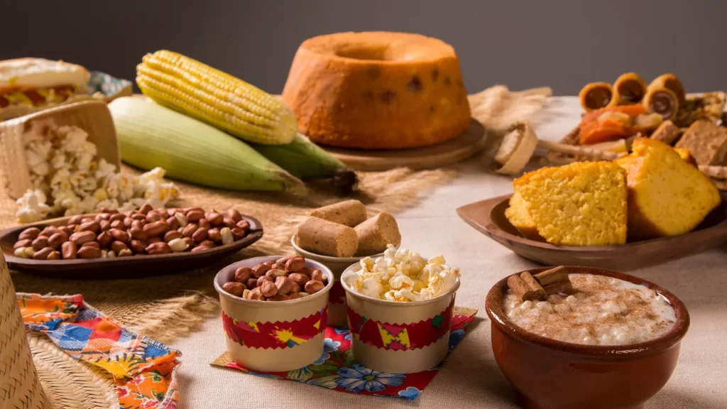 Mesa com comidas típicas da culinária caipira, incluindo bolo de milho, pamonha, canjica, amendoim, pipoca e espigas de milho, decorada com elementos de festa junina.